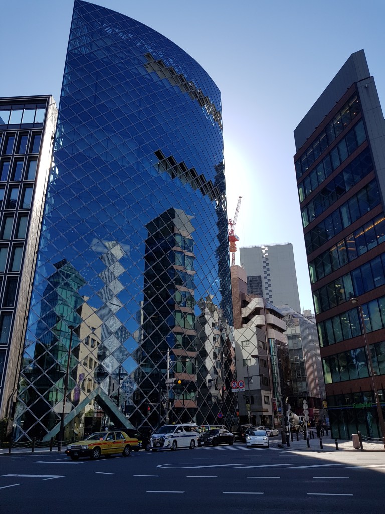 Moderne Architektur eines blauen Glasgebäudes mit ungewöhnlicher spitzer Form in einer belebten Stadtstraße, umgeben von Taxis und weiteren Hochhäusern bei klarem Himmel