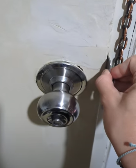 Close-up of a hand inserting a travel door lock into a door strike plate, showcasing a stainless steel door knob and a chain lock.