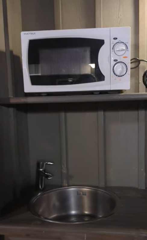 A shelf featuring a microwave and a stainless steel sink, set against a wooden wall.